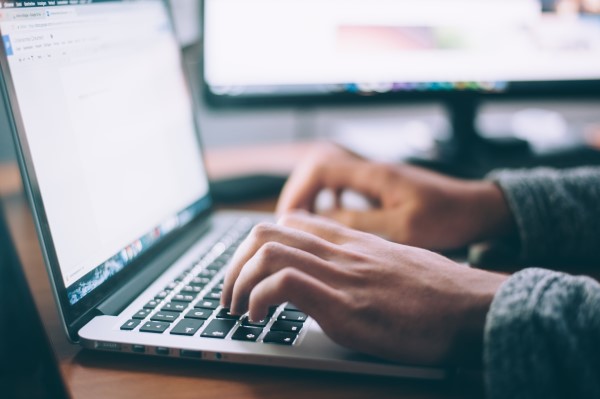 Person typing on a laptop keyboard with an external monitor in the background